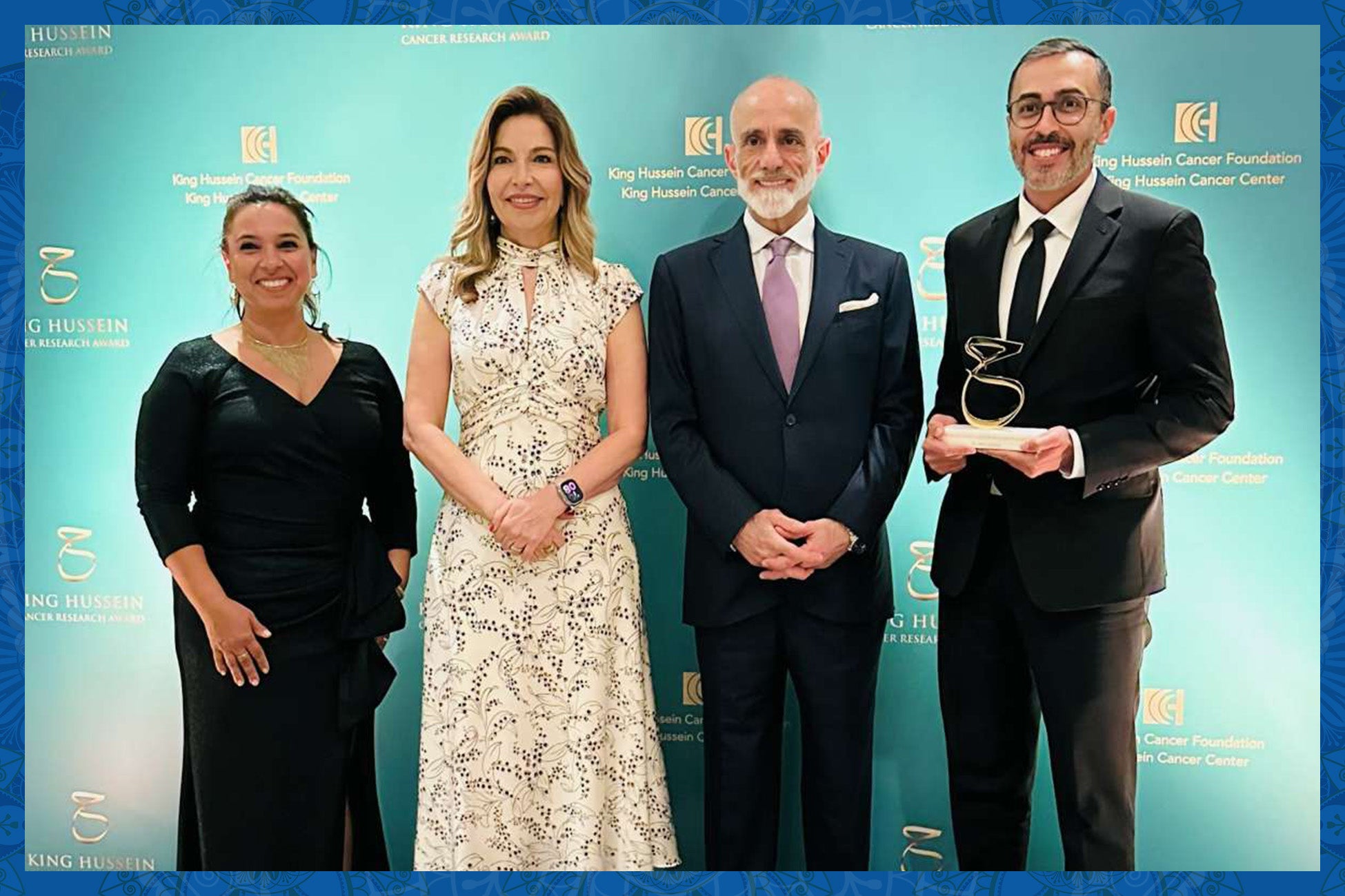 Four people stand in front of a teal backdrop. Two of them are women and two of them are men. They are all dressed in formal wear. The man on the far right is holding a trophy. 